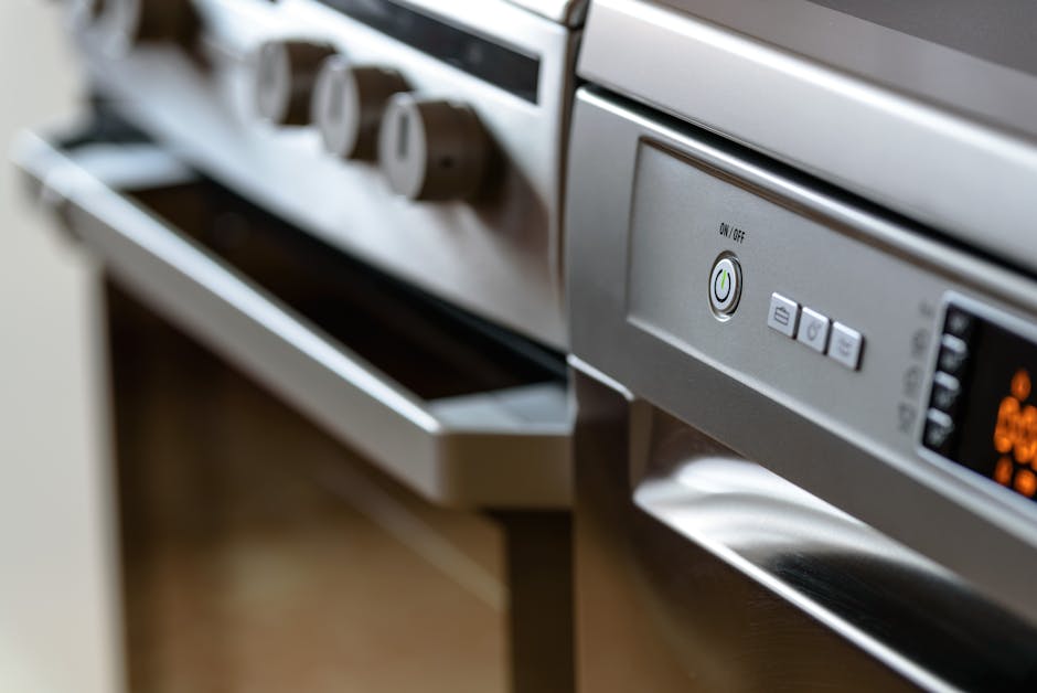 A woman with red hair, wearing a white t-shirt and pink apron, is wiping a black glass ceramic cooktop on a white kitchen countertop with a cloth. The kitchen features white cabinets and a grey backsp