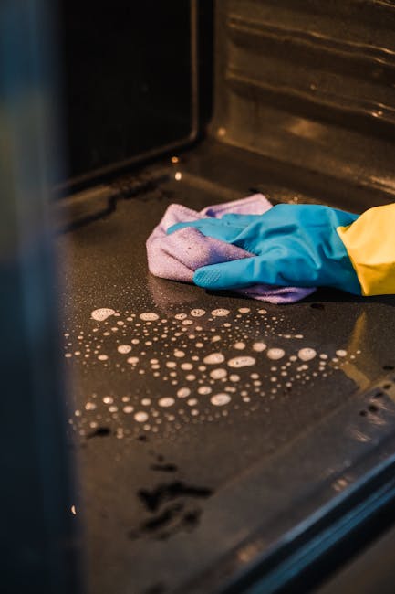 Close-up of a black, non-stick oven surface being cleaned, featuring soap suds and water droplets. A person wearing yellow and blue cleaning gloves holds a pink and purple microfiber cloth, wiping the oven's interior. The oven's walls are dark with visible signs of previous use, while the surface appears wet and partially cleaned, indicating the ongoing deep cleaning process. Bright lighting illuminates the area, highlighting the shiny, reflective surface and freshly applied cleaning solution. This image exemplifies detailed surface cleaning, emphasizing hygiene and maintenance, with the focus on the oven's interior as part of domestic cleaning services provided by Oven Cleaning Kingston, specializing in oven surface sanitisation and deep cleaning. The background and surrounding elements are minimal, keeping the emphasis on the cleaning action and materials used.
