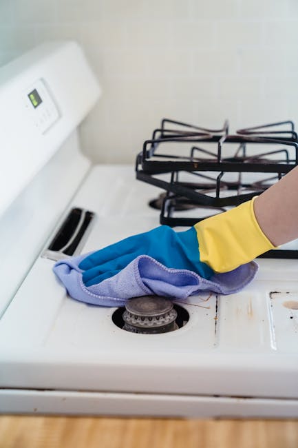A close-up view of a white gas stovetop in a kitchen, with a person's hand wearing a yellow rubber glove and holding a purple cleaning cloth, thoroughly wiping the surface around the burner. The stove features metallic grates over two burners, with one burner visibly being cleaned. The background shows a beige tiled wall, and the stove surface appears clean and slightly shiny, indicating recent or ongoing surface cleaning and sanitisation, consistent with domestic cleaning services provided by Oven Cleaning Kingston for the Bentall Centre oven cleaning services Kingston, Kingston page.