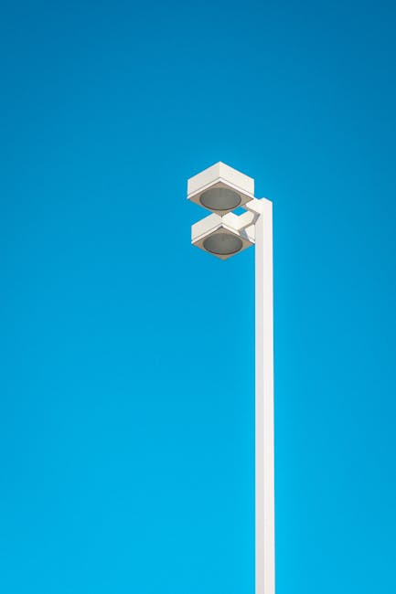 A tall, white street lighting pole stands prominently against a clear blue sky, featuring a modern rectangular lamp head with multiple circular light fixtures. The pole extends vertically from the bot