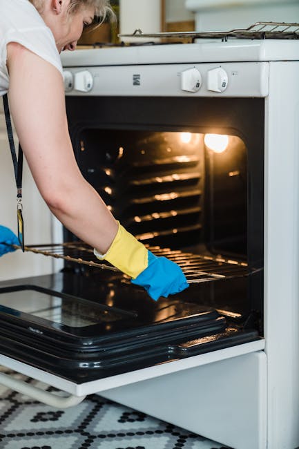 A person wearing yellow and blue rubber gloves is cleaning the inside of a black oven with a pink cloth, focusing on the oven's surface which has some soap or cleaning solution residues visible. The o